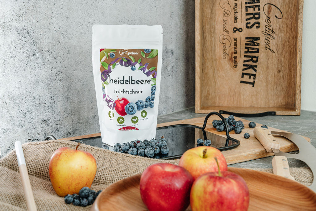 Fruchtschnurtüte Geschmacksrichtung Heidelbeere und im Vordergrund verschiedene Früchte alles auf einem Schneidbrett