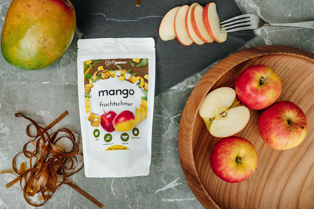 36g Tüte Fruchtschnüre Mango mit umliegenden Früchten: Apfel in Schale und auf Schieferunterlage, Mango und ausgepackte Fruchtschnüre auf grauem Tisch als Unterlage