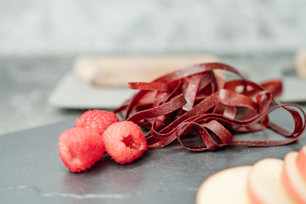 Auf einer Schieferplatte liegen Himbeeren, Fruchtschlangen Sorte Himbeere sowie Apfelschnitze, im Hintergrund liegt ein Wiegemesser