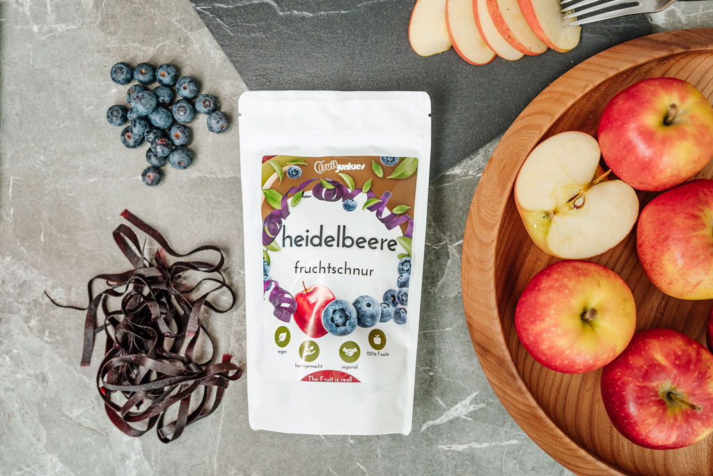 Heidelbeere Fruchtschnurtüte umliegend ganze und aufgeschnittene Äpfel, ausgepackte Äpfel, Heidelbeeren und eine Schieferplatte auf grauer Tischunterlage