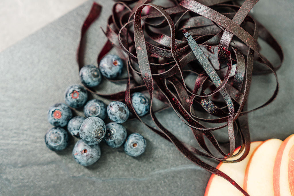 Auf einer Schieferplatte liegen Heidelbeeren, Fruchtschlangen Sorte Heidelbeere sowie Apfelschnitze