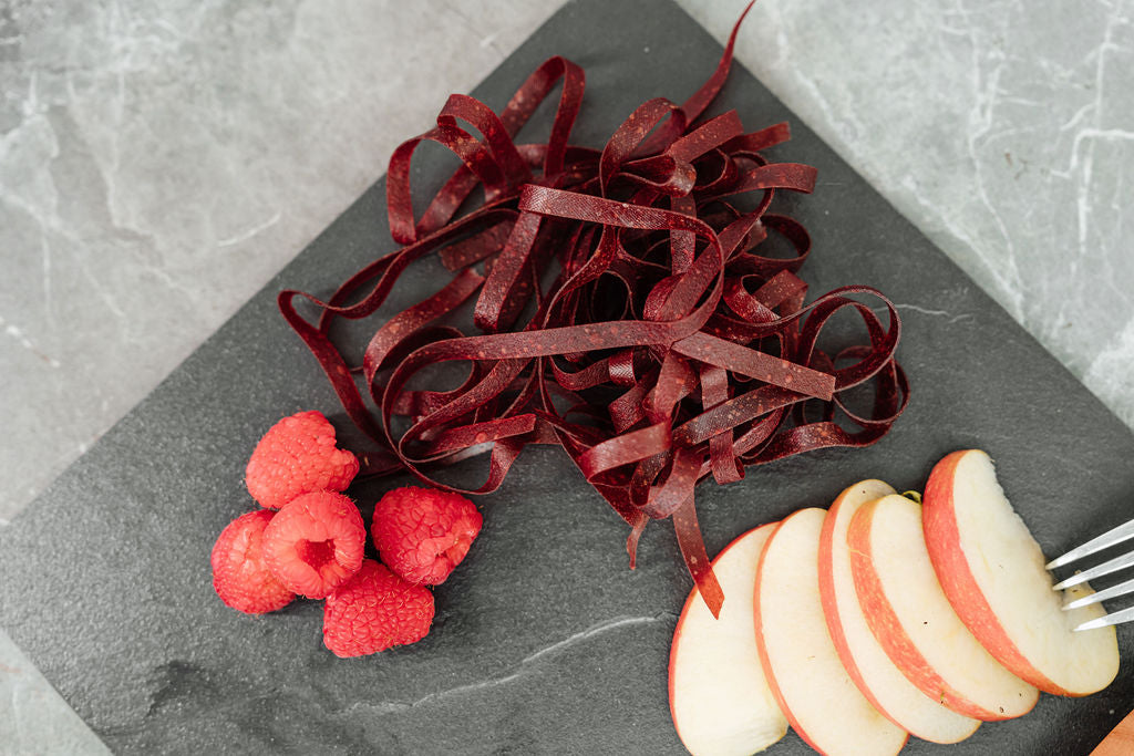 Auf einer Schieferplatte liegen Himbeeren, Fruchtschlangen Sorte Himbeeren sowie Apfelschnitze und eine Gabel