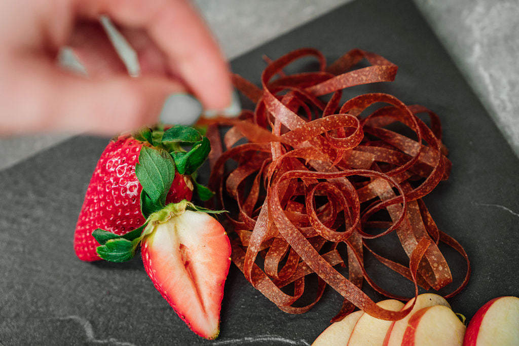 Auf einer Schieferplatte liegen Erdbeeren, Fruchtschlangen Sorte Erdbeere sowie Apfelschnitze und eine Hand die die Fruchtschlangen nimmt