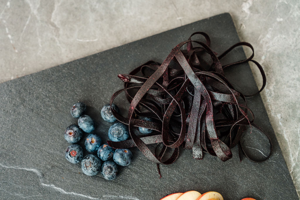 Auf einer Schieferplatte liegen Heidelbeeren, Fruchtschlangen Sorte Heidelbeere sowie Apfelschnitze