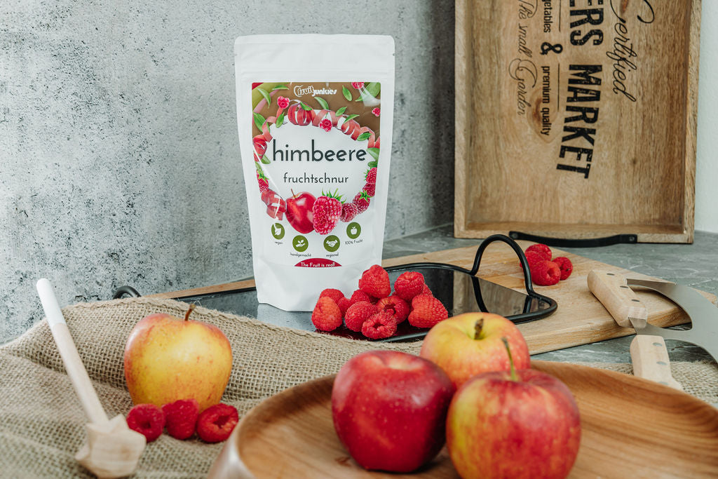 Fruchtschnurtüte Geschmacksrichtung Himbeere und im Vordergrund verschiedene Früchte alles auf einem Schneidbrett