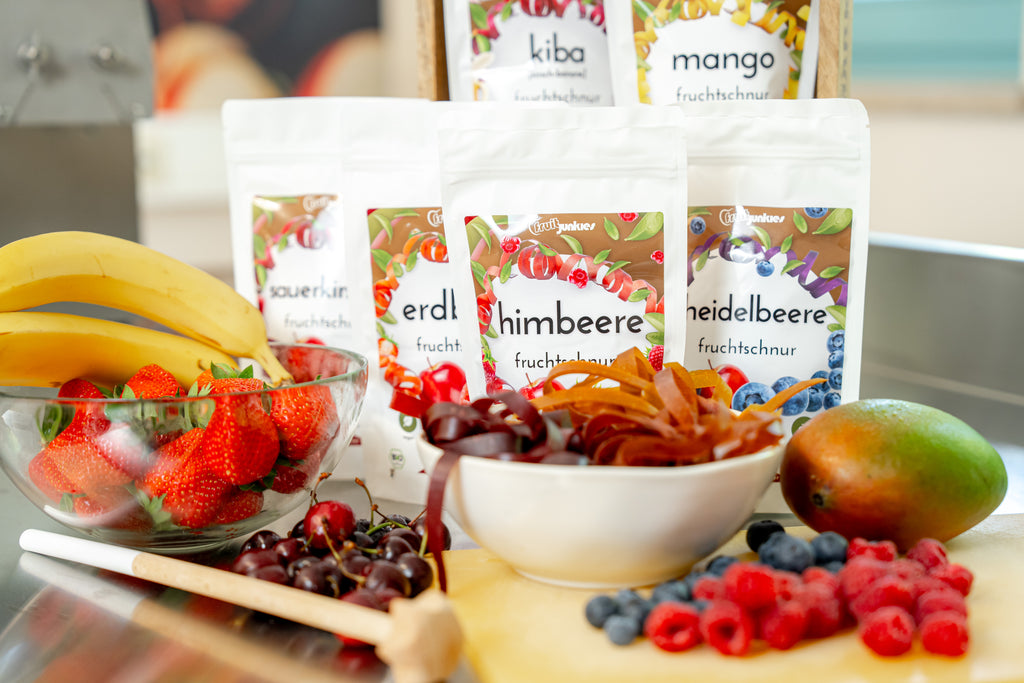 36g Tüten verschiedener Geschmacksrichtungen von Fruchtschnüren, Fruchtschnüre in Schale und Obst vor den Tüten sowie Kochutensilien