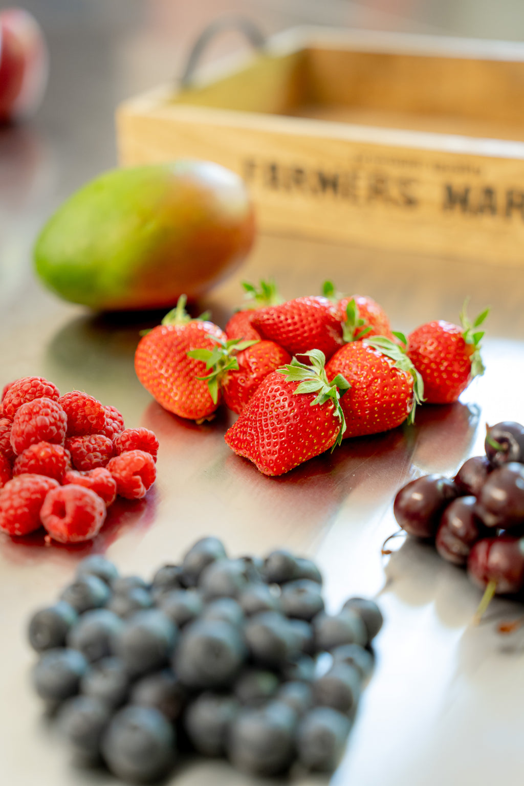 Auf Edelstahltisch liegende Mango, Erdbeere, Sauerkirschen, Himbeeren und Heidelbeeren. Im Hintergrund Holztablett als Deko