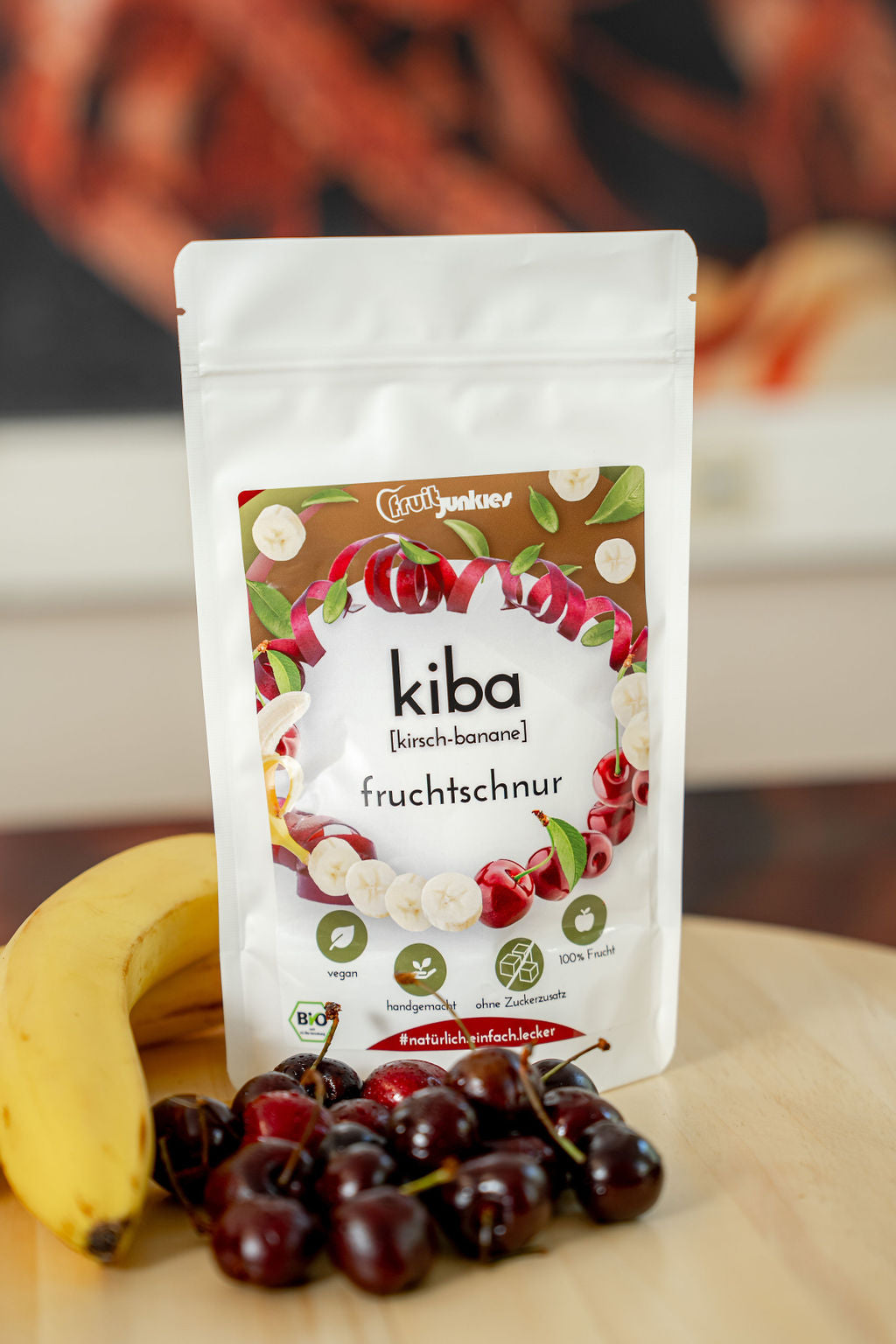 36g Fruchtschnurtüte Sorte KiBa auf einem Holzteller mit einer Banane und Sauerkirsche